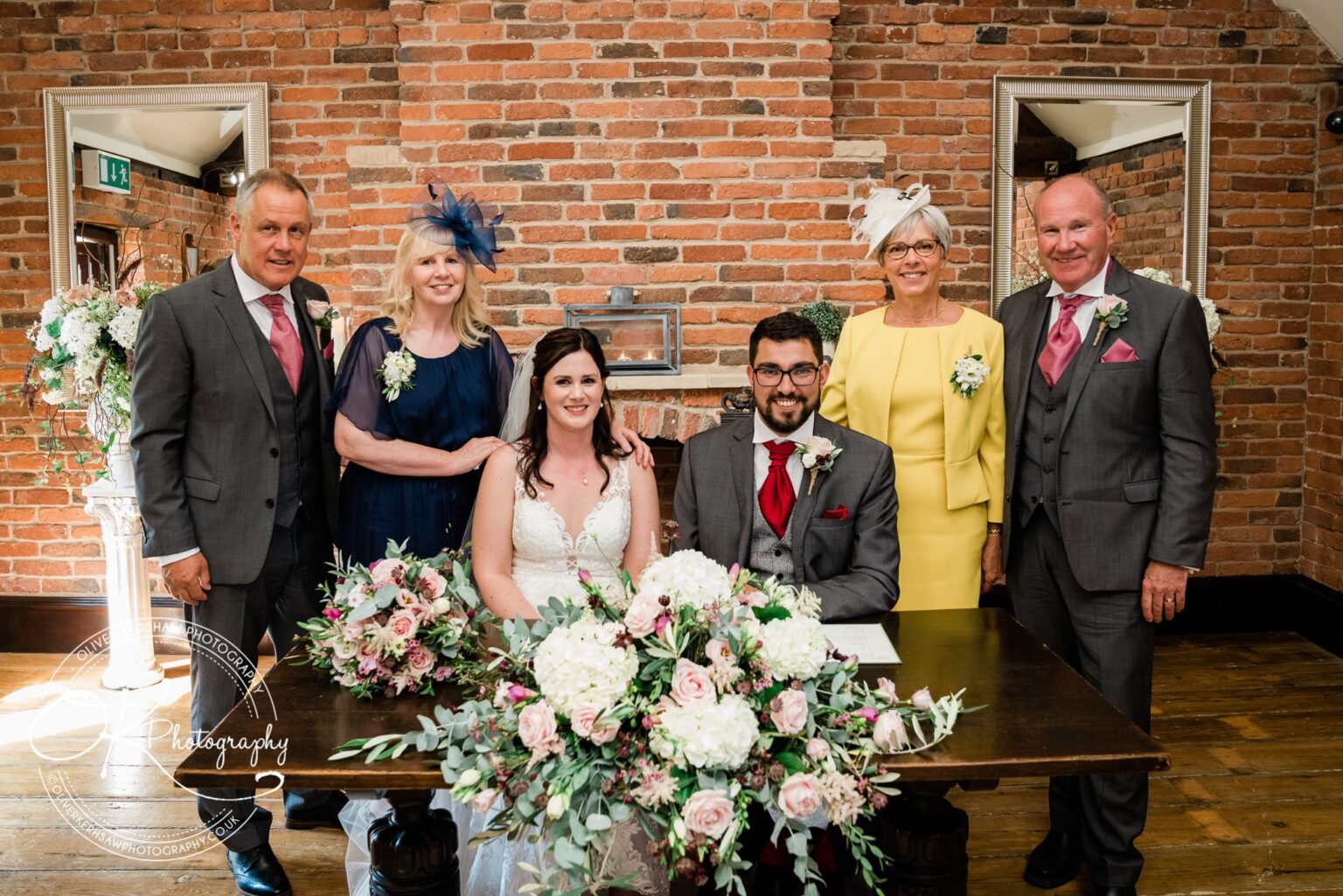 A bride and groom seated at a table with floral arrangements, surrounded by four elegantly dressed adults, posing for a wedding photograph indoors with a brick wall background.