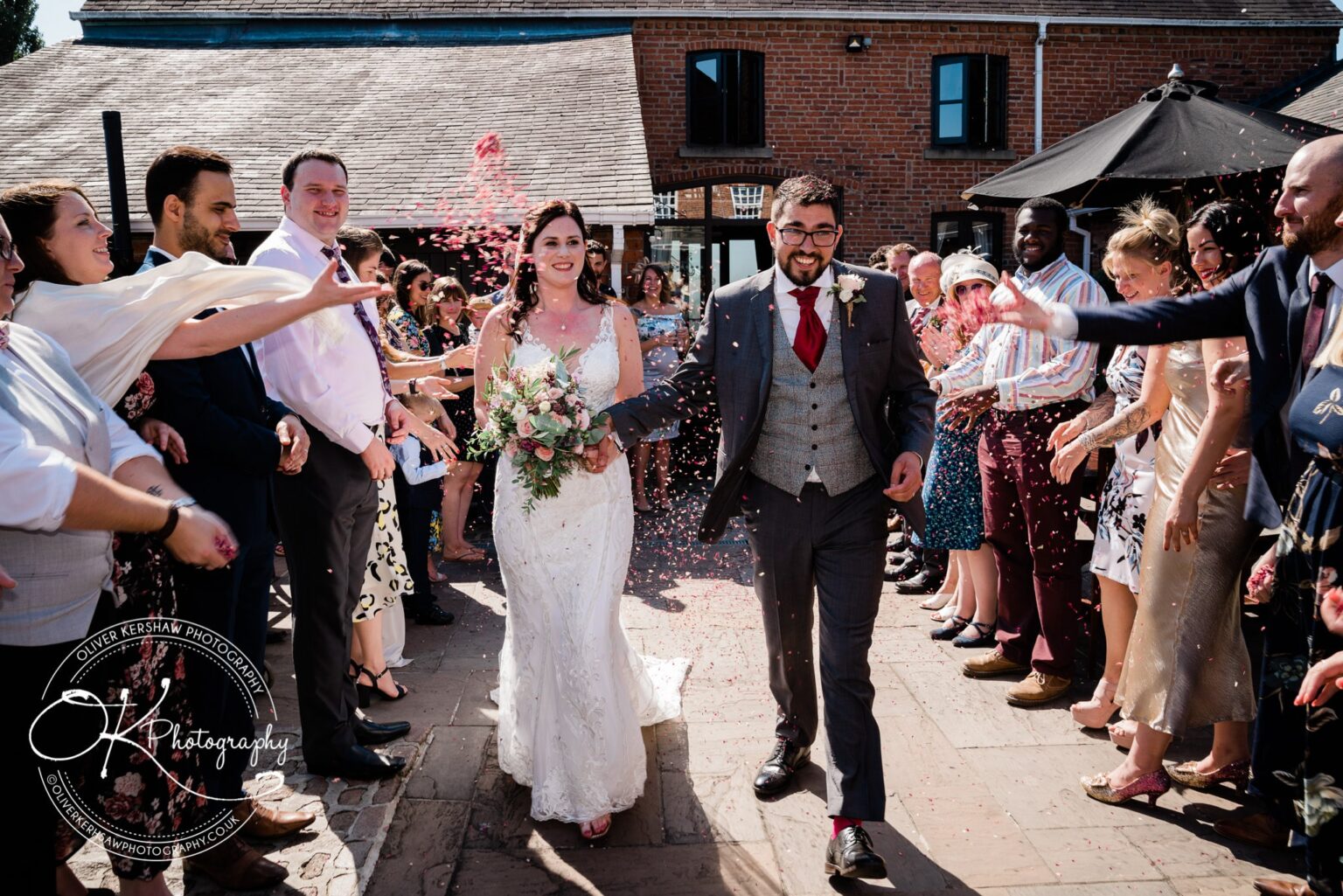 Bride and groom walking through a crowd of guests throwing confetti outside a brick building.
