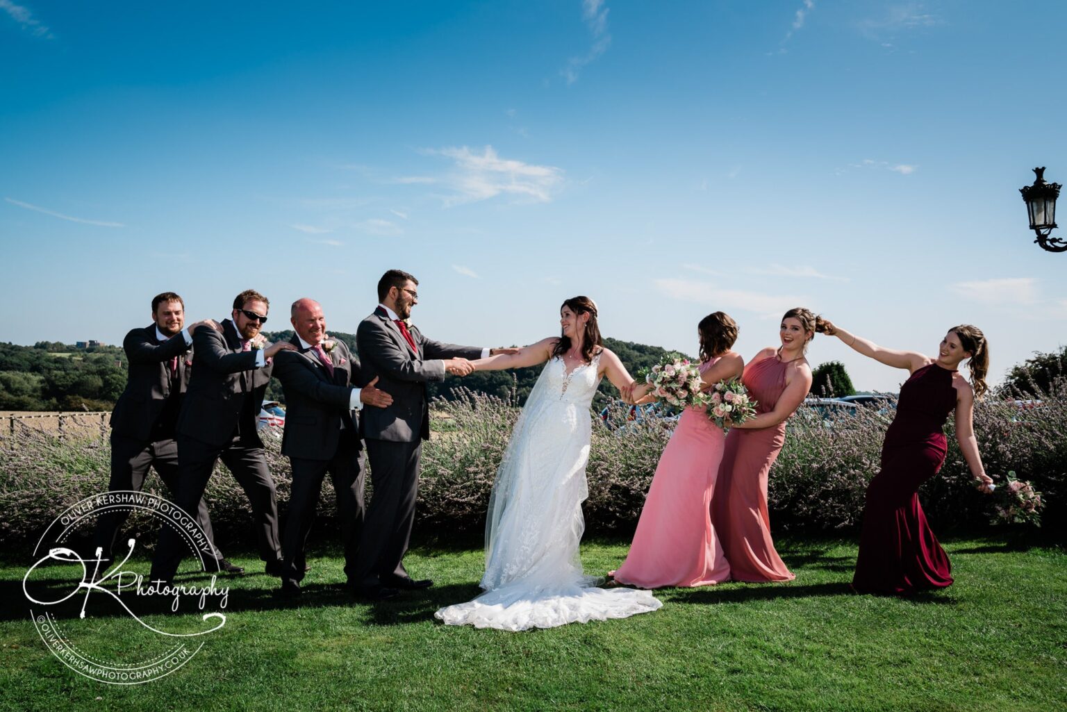 Wedding party playfully posing in a tug-of-war style at Swancar Farm, with groomsmen on one side and bridesmaids on the other, under a clear blue sky.
