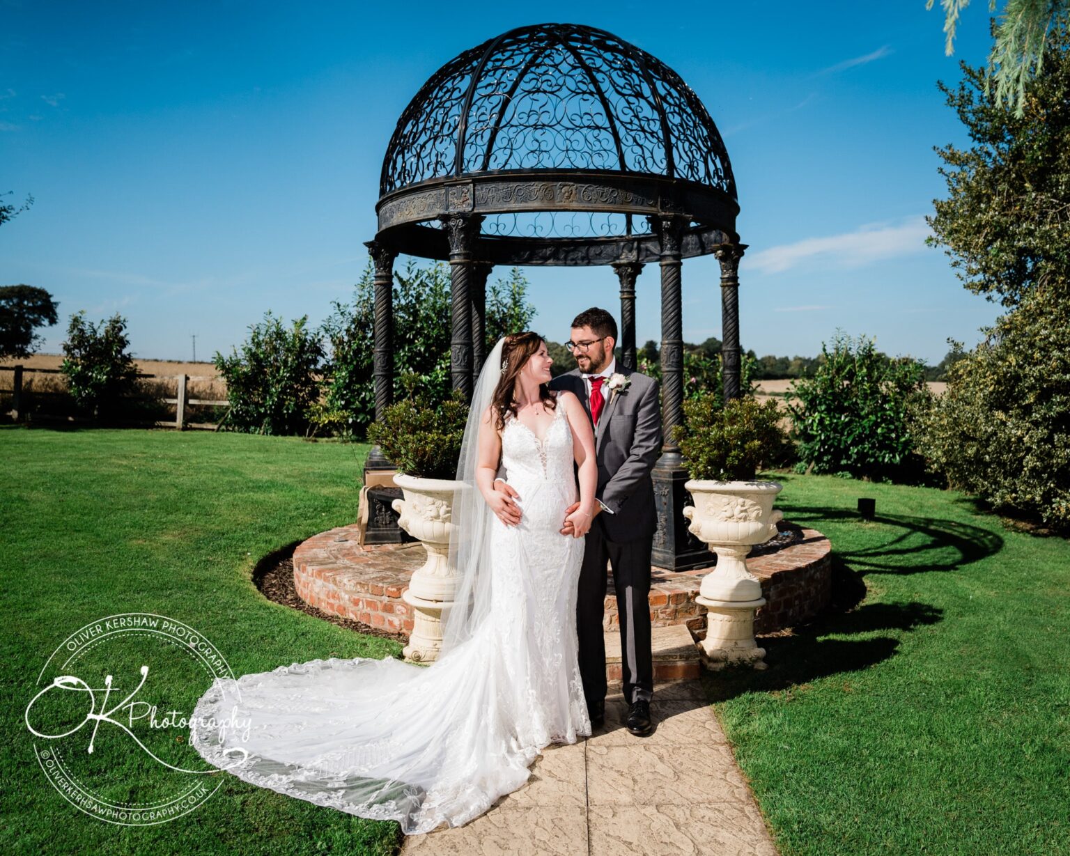 A bride and groom pose together in a garden under a decorative metal gazebo at Swancar Farm, photographed by Oliver Kershaw Photography.