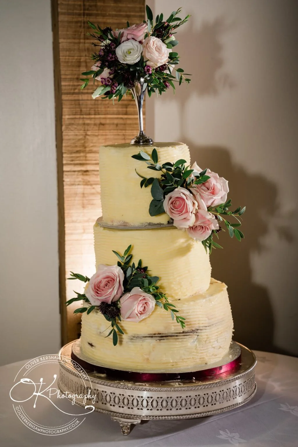 Three-tiered rustic wedding cake decorated with pink and white flowers, placed on a silver cake stand.