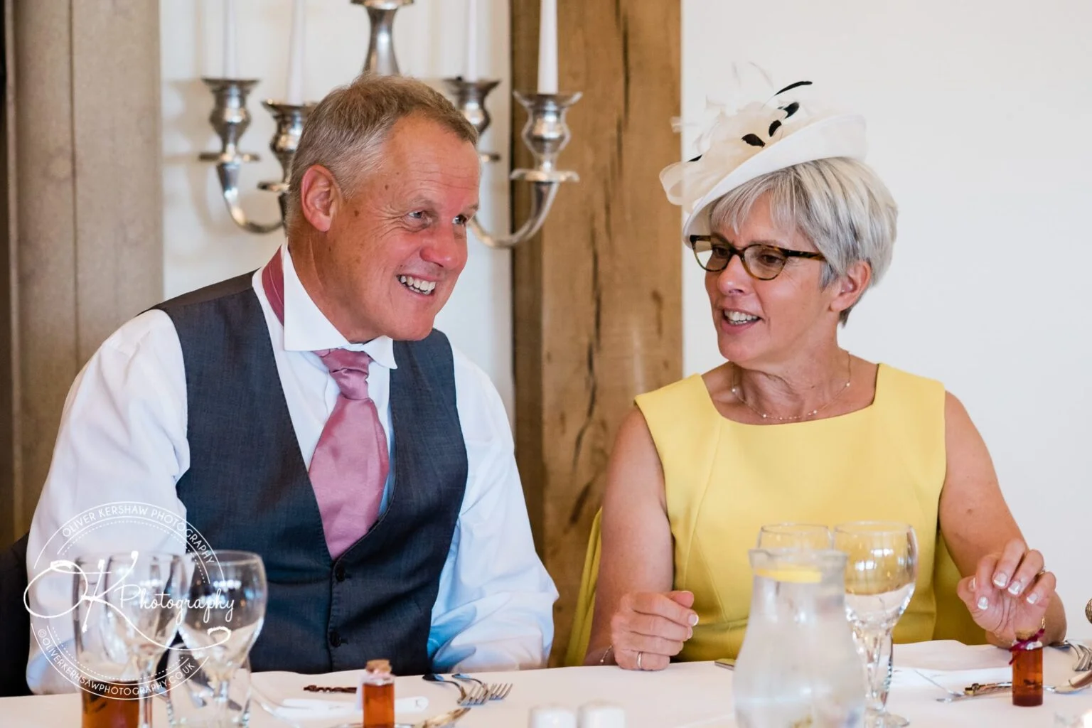 A man in a grey waistcoat and pink tie smiles while conversing with a woman in a yellow dress and white hat at a dining table.