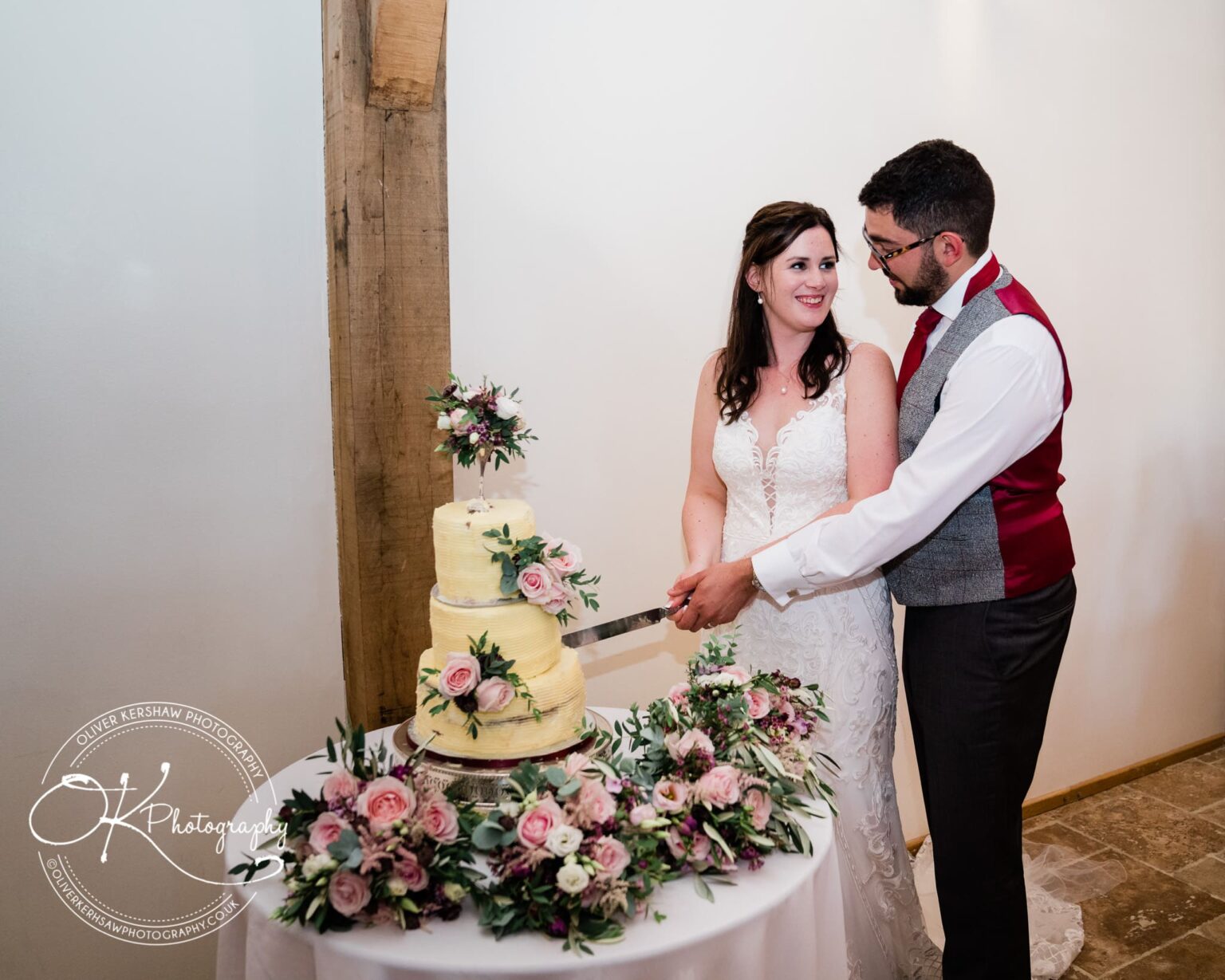 A bride and groom cutting a tiered wedding cake adorned with flowers, inside a rustic venue.