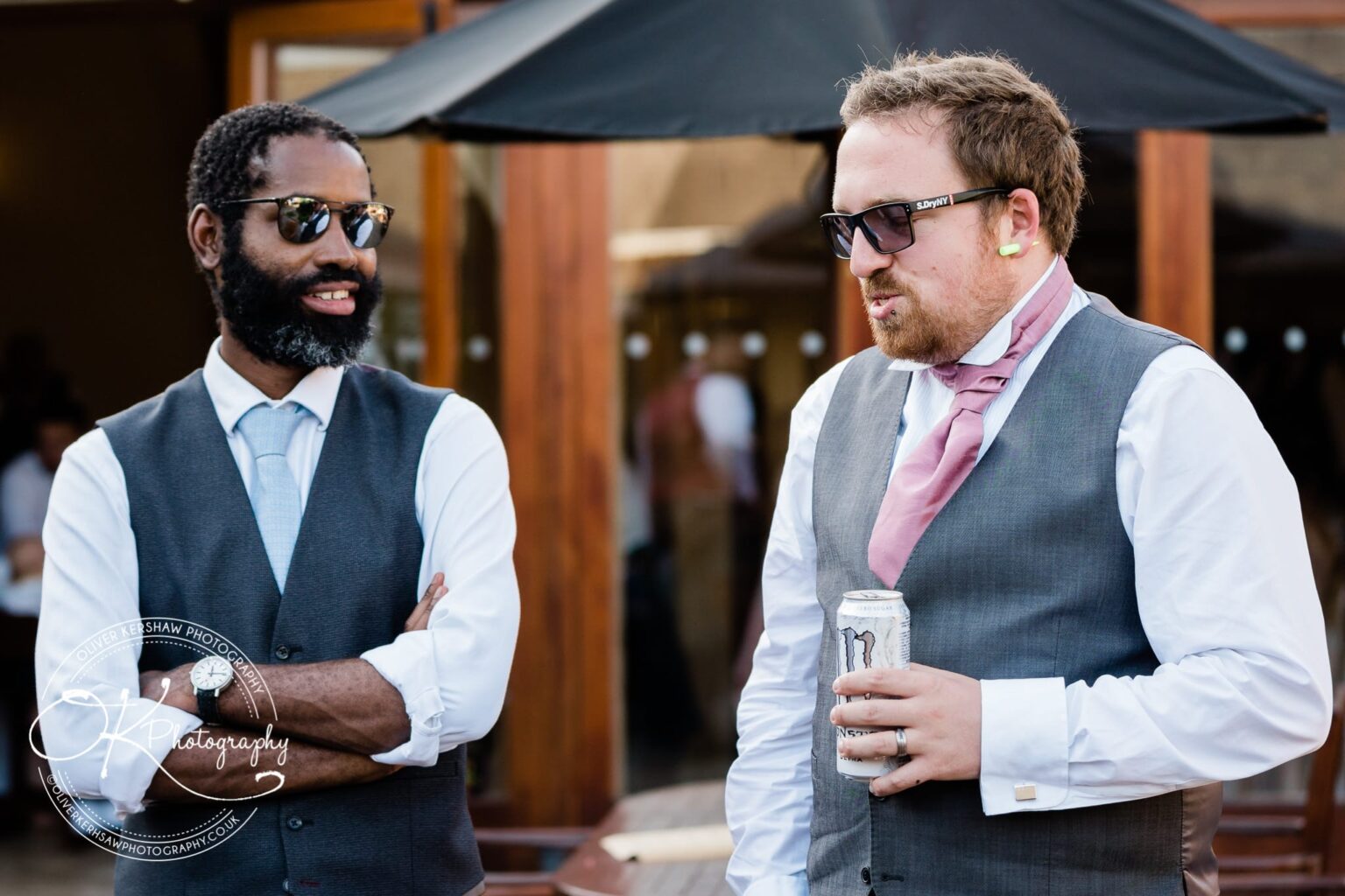 Two men wearing formal attire and sunglasses, engaged in conversation. One holds a drink can. One of them has a beard and wears a vest, while the other wears a skewed tie and holds an energy drink.
