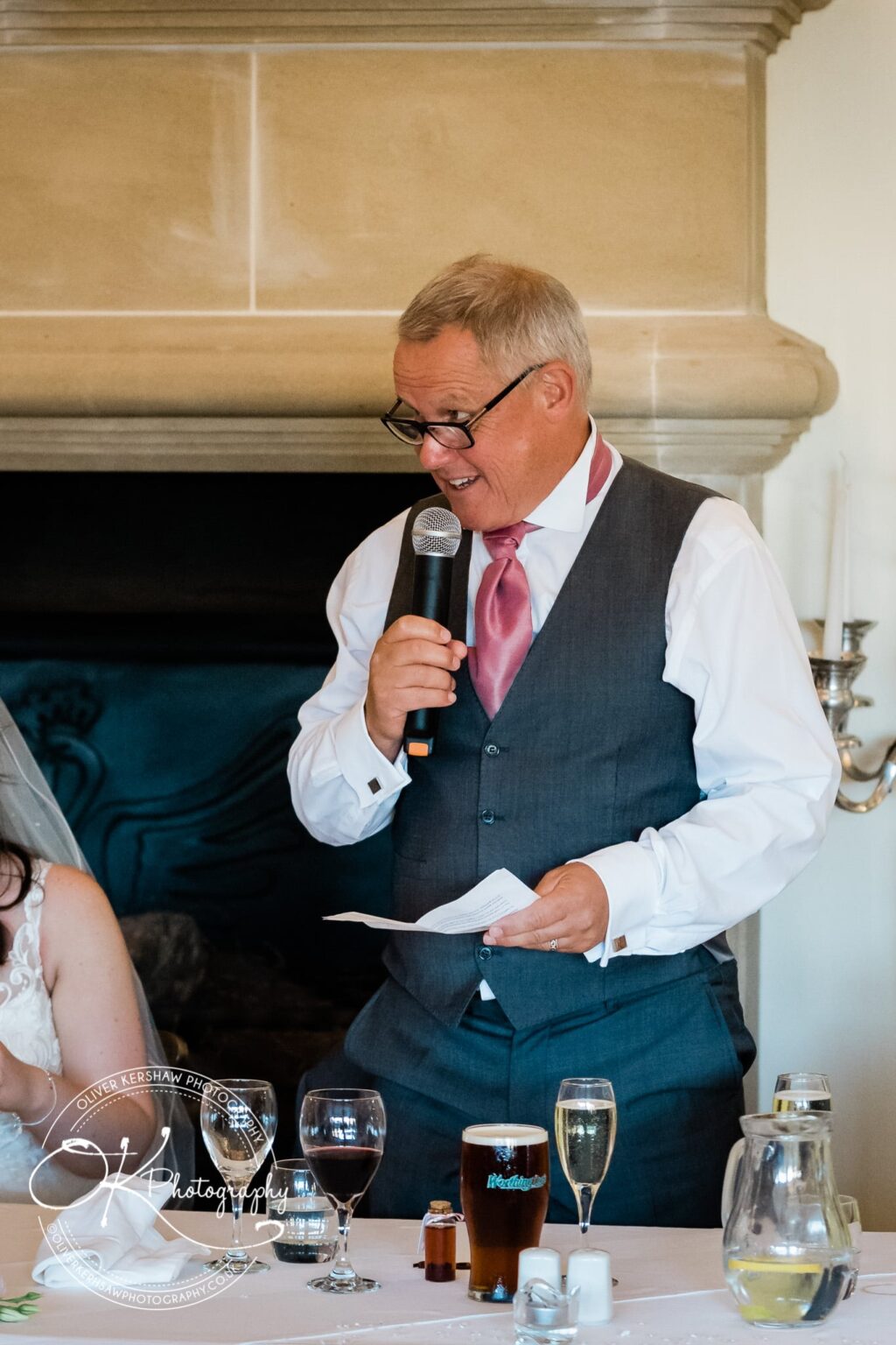 An older man in a grey vest and pink tie giving a speech with a microphone at a wedding reception.