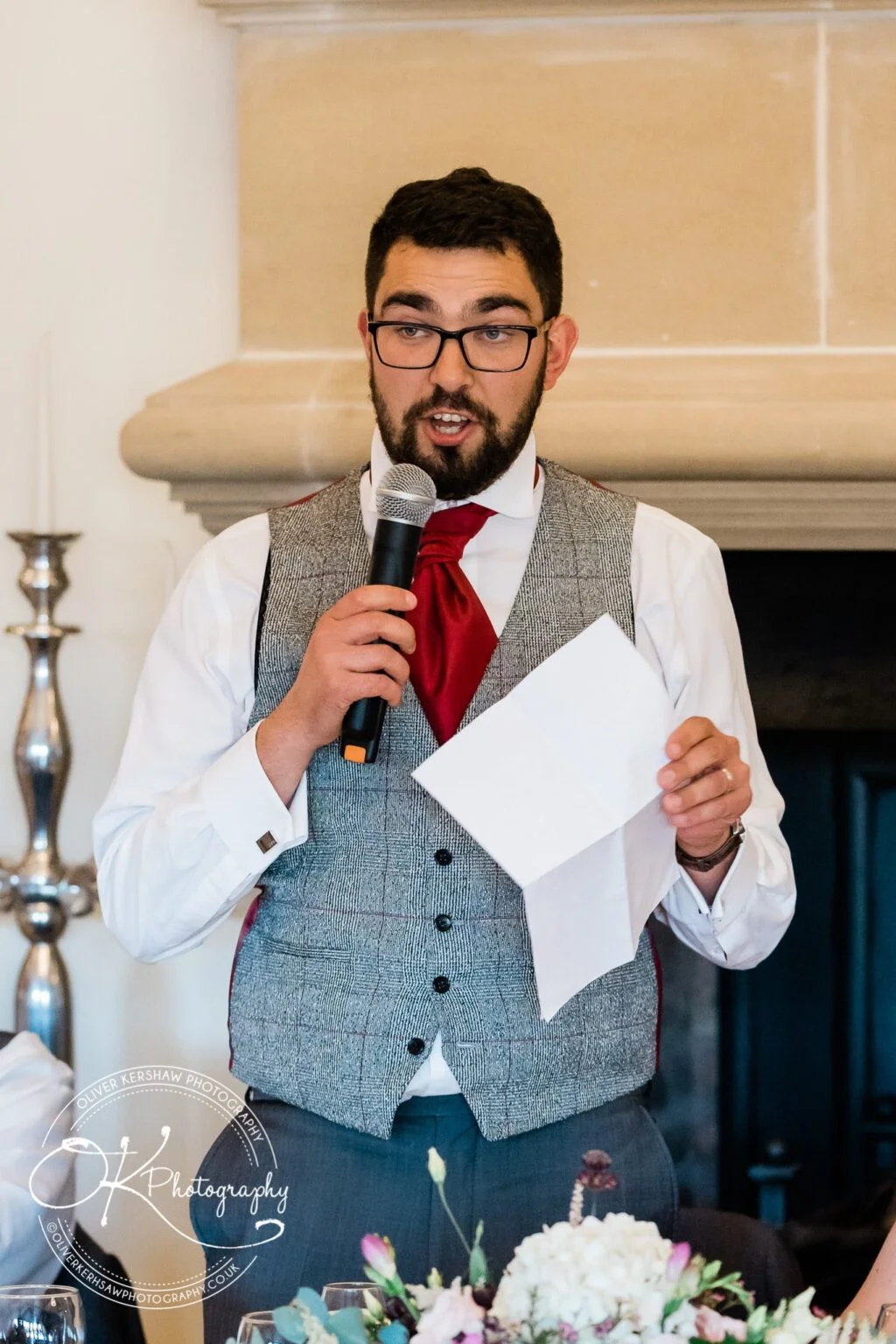 A man wearing glasses and a grey waistcoat with a red tie gives a speech while holding a microphone and a piece of paper, standing in front of a decorated setting at a wedding reception.