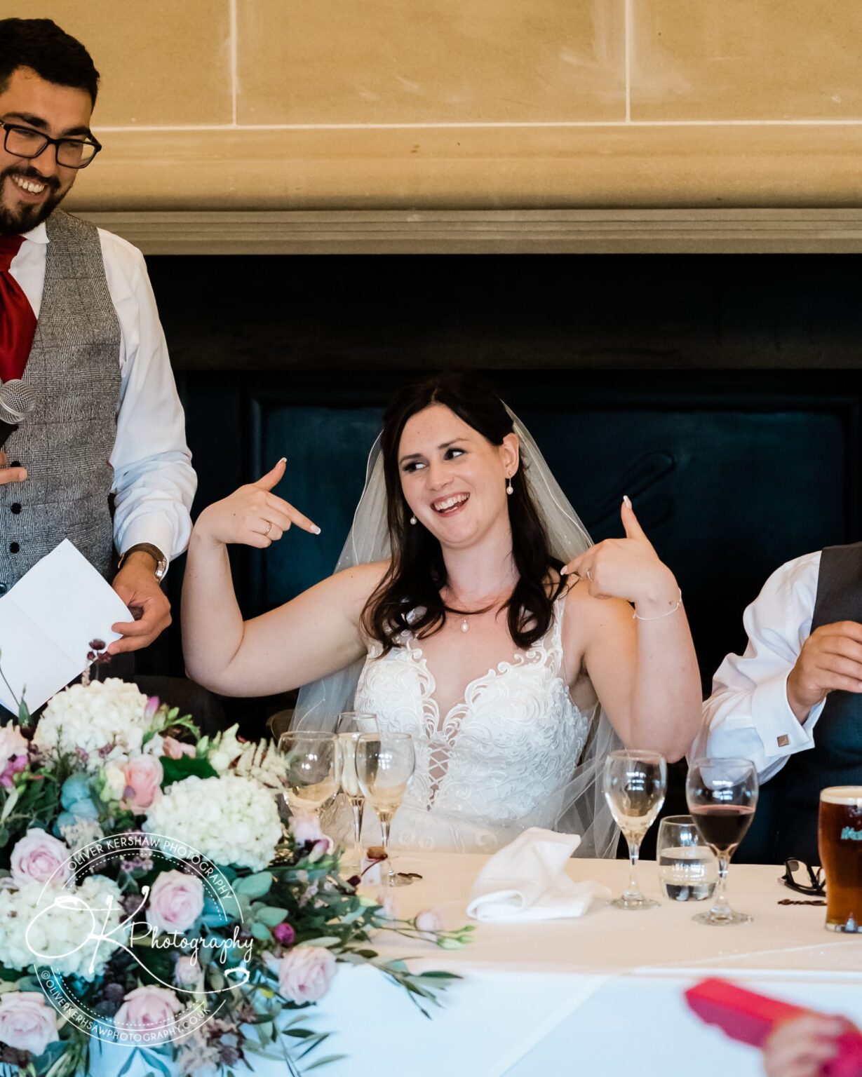 Bride in a white dress and veil sitting at a table, smiling and pointing at herself, with a man standing next to her holding a microphone.