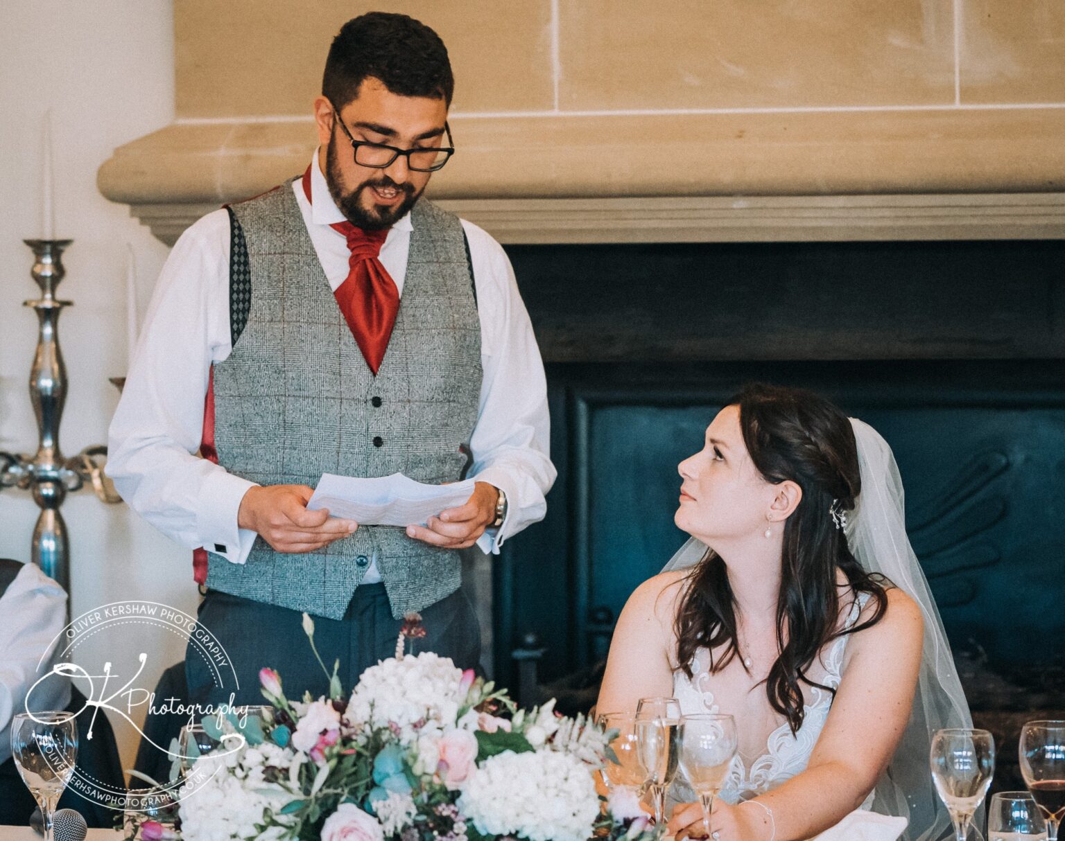 Man in a suit and red cravat giving a speech, while a woman in a wedding dress and veil looks up at him, with a floral arrangement and empty wine glasses on the table.