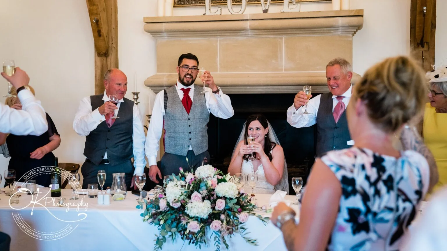 Wedding party toasting with drinks around a table, with a flower arrangement and the word "LOVE" visible on a mantle in the background.