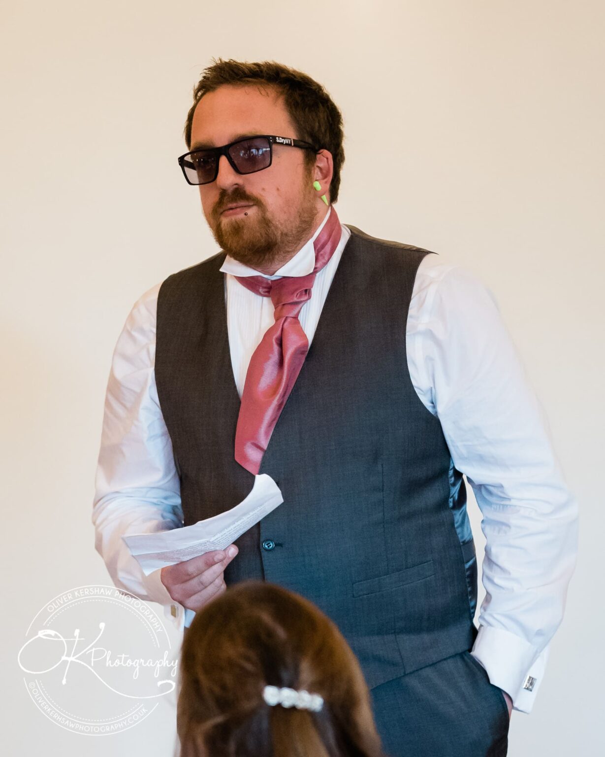 Man in a waistcoat and pink cravat holding a paper, wearing sunglasses and earplugs, giving a speech at a wedding.