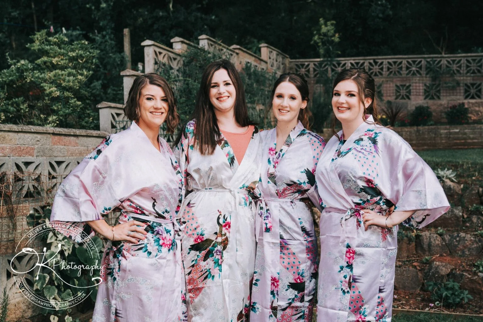 Four women wearing matching floral satin robes pose together outside.