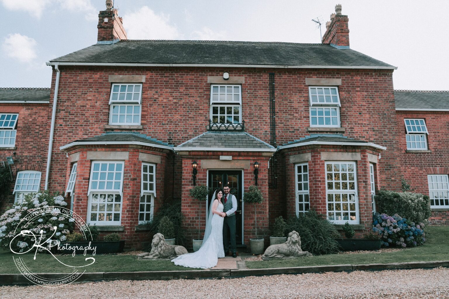 A newlywed couple stands in front of a large red-brick house with bay windows at Swancar Farm.