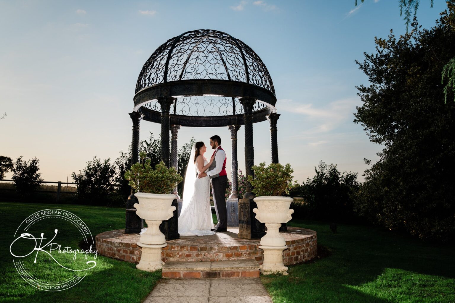A bride and groom standing under an ornate, wrought-iron gazebo in a garden during their wedding ceremony at sunset.