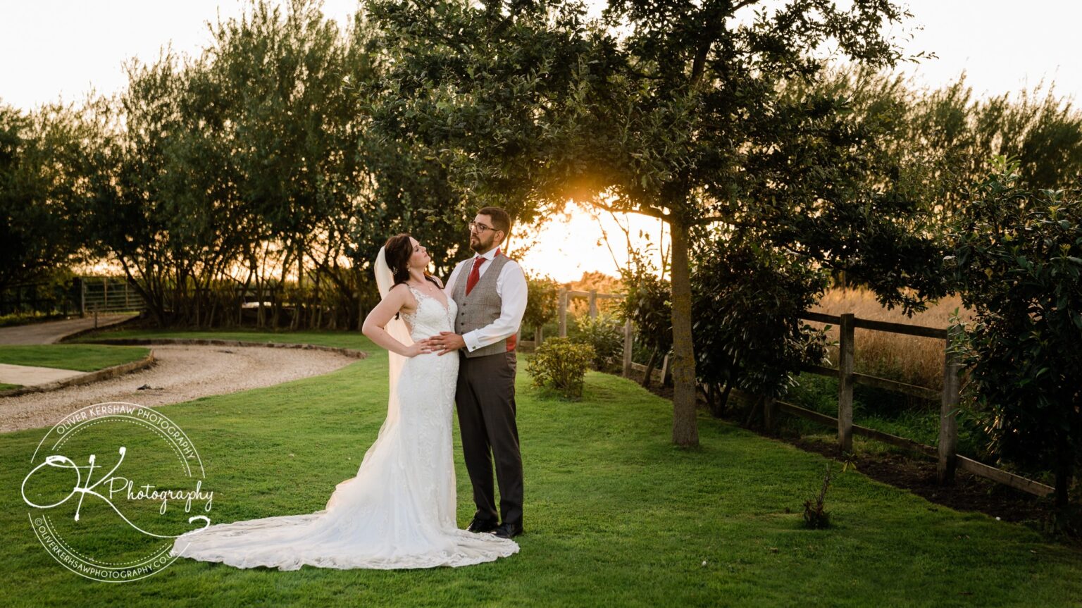 A bride and groom standing on grass under a tree at sunset, captured by OK Photography at Swancar Farm.