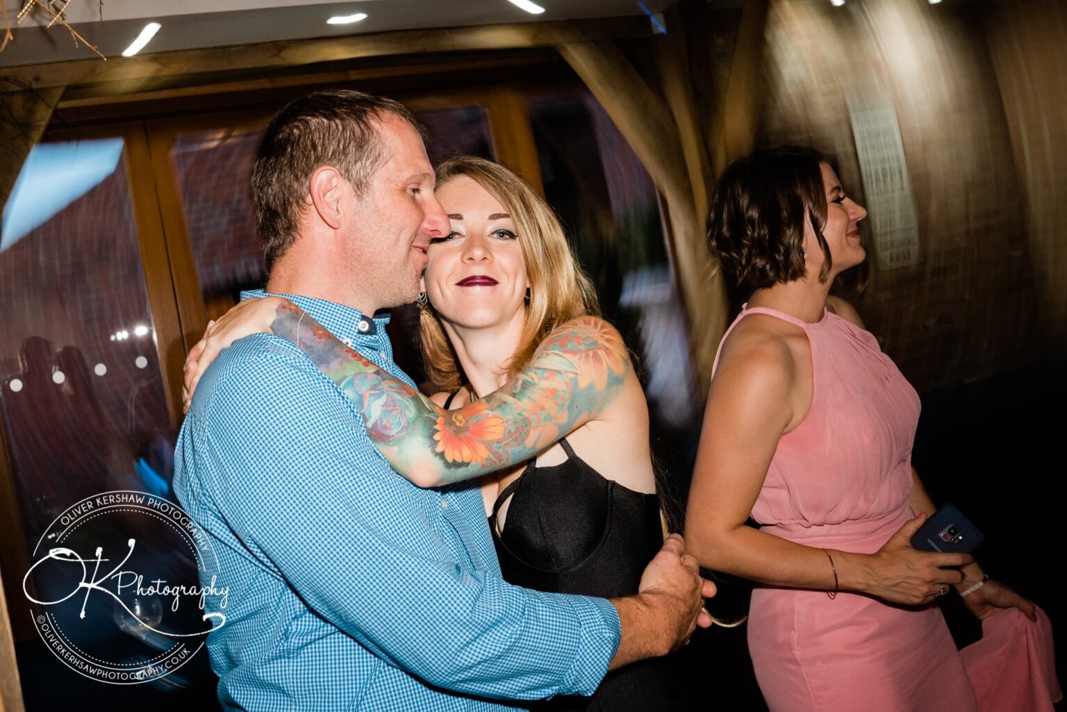 A couple embraces at a wedding reception, with the woman in a black dress showing colorful tattoos on her arms, while another woman in a pink dress stands nearby, holding a mobile phone.