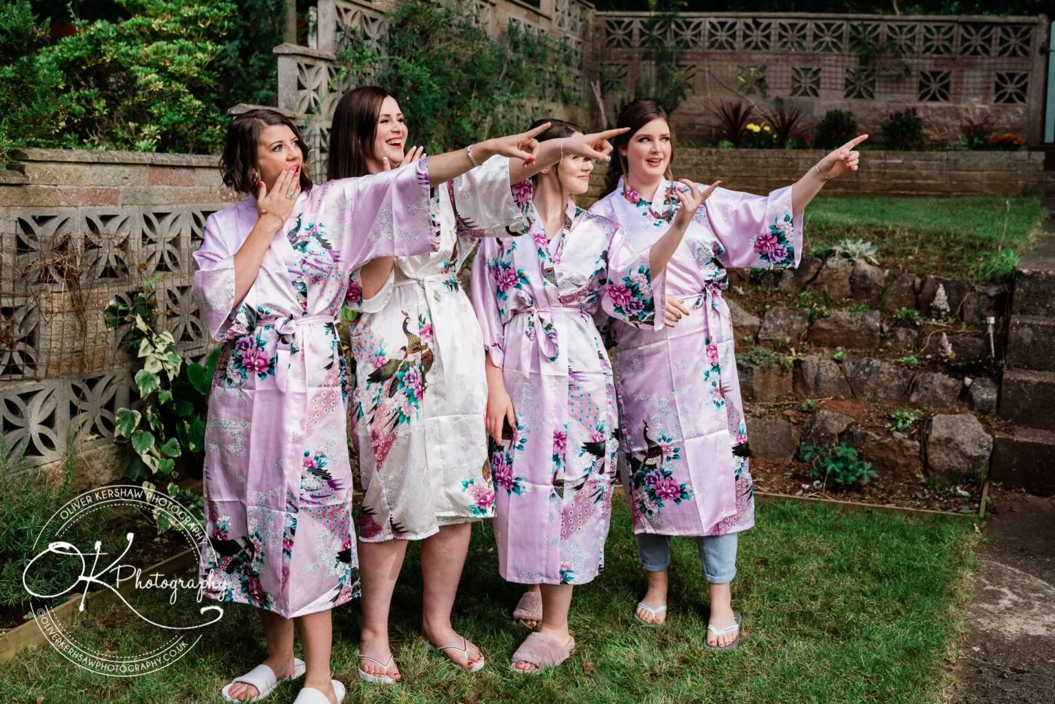 Four women in floral robes and slippers pointing in the same direction while standing outdoors on grass.