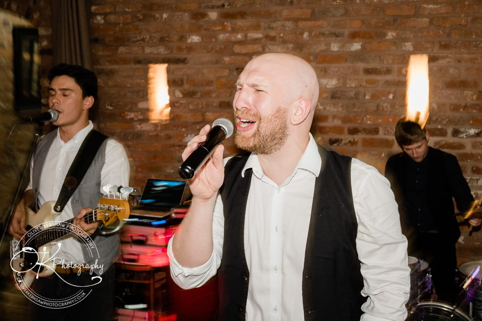 A man is singing into a microphone while another man plays a guitar in the background. The setting features an exposed brick wall and ambient lighting.