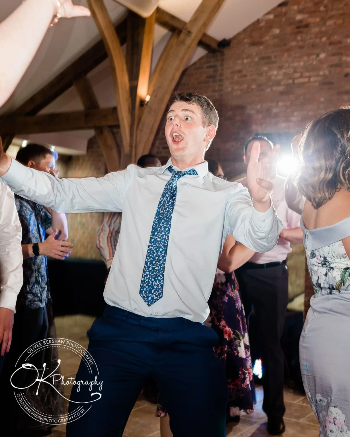 A man in a white shirt with a floral tie dances enthusiastically at an event, surrounded by other people.