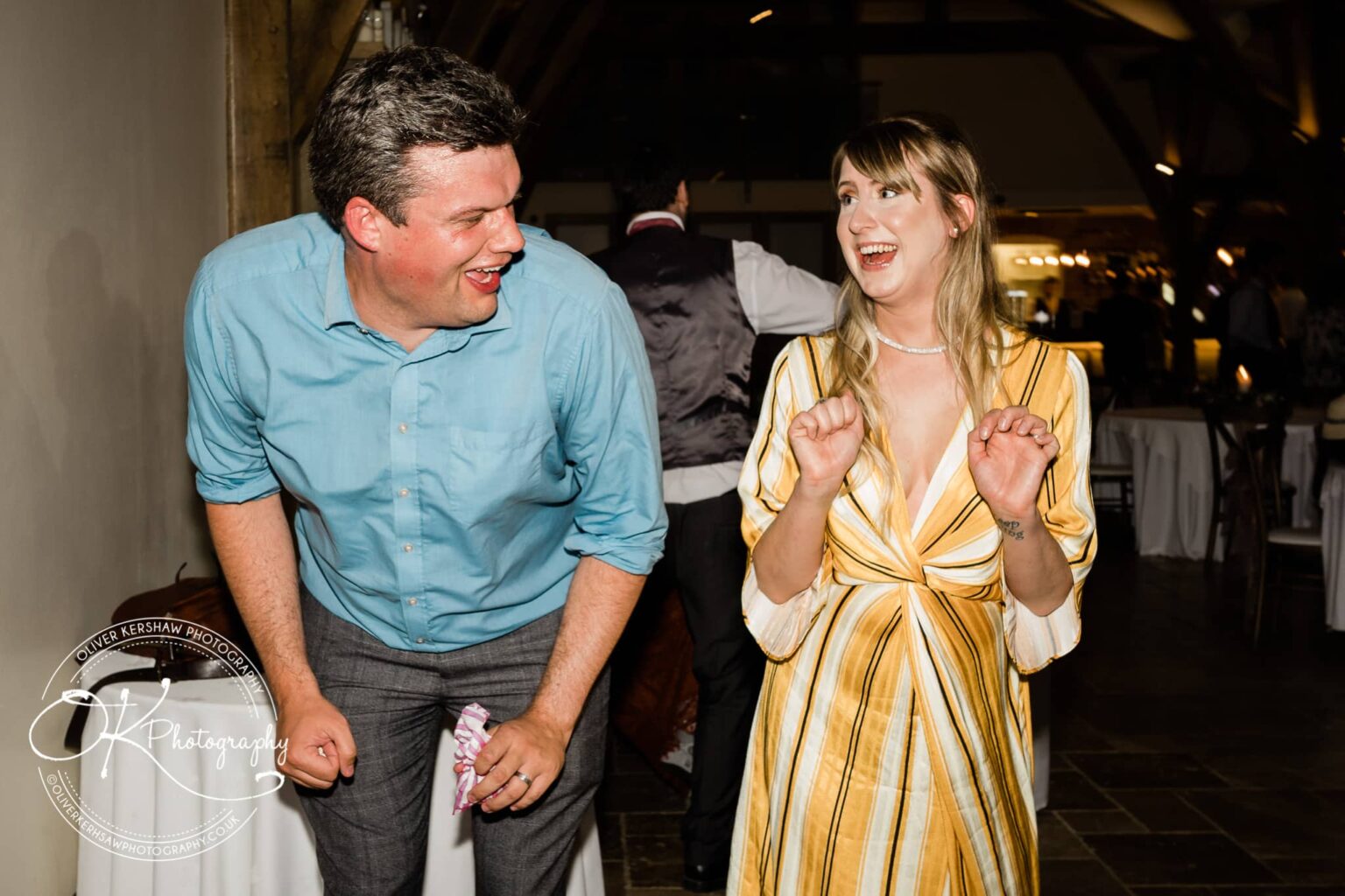 A man in a blue shirt and a woman in a yellow striped dress dance and laugh at a wedding reception.