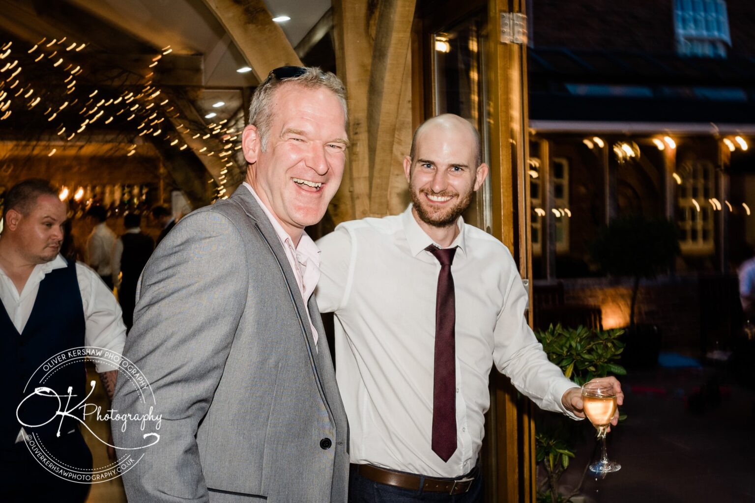 Two men smiling at a wedding reception, one holding a glass of wine, with warm lighting and fairy lights in the background.