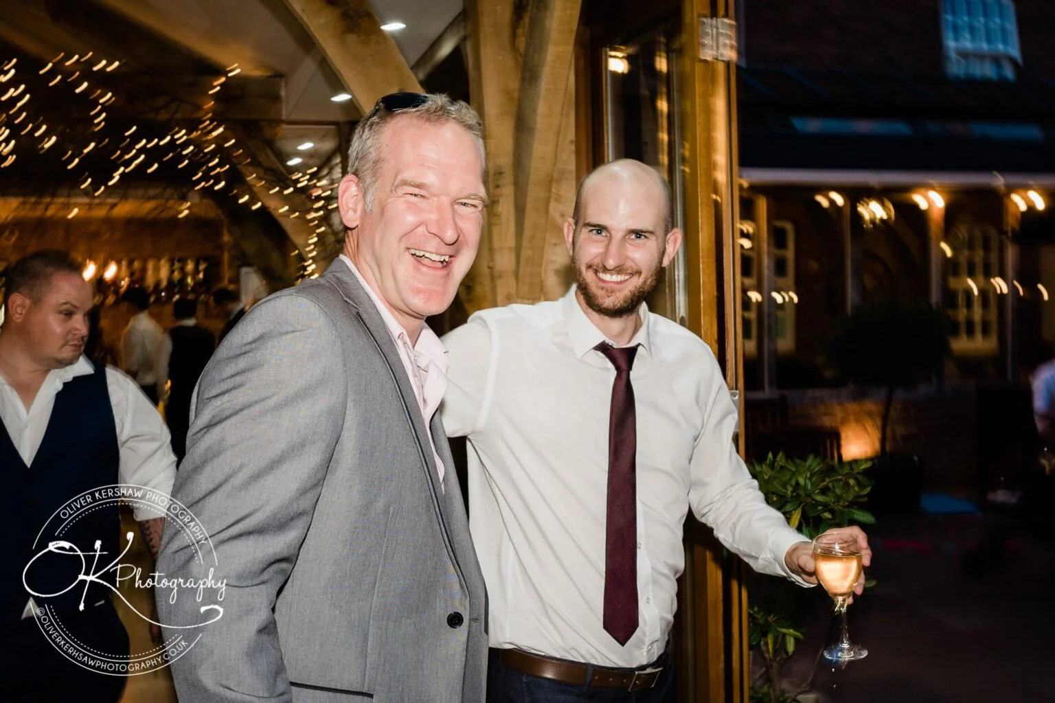 Two men smiling at a wedding reception, one holding a glass of wine, with warm lighting and fairy lights in the background.