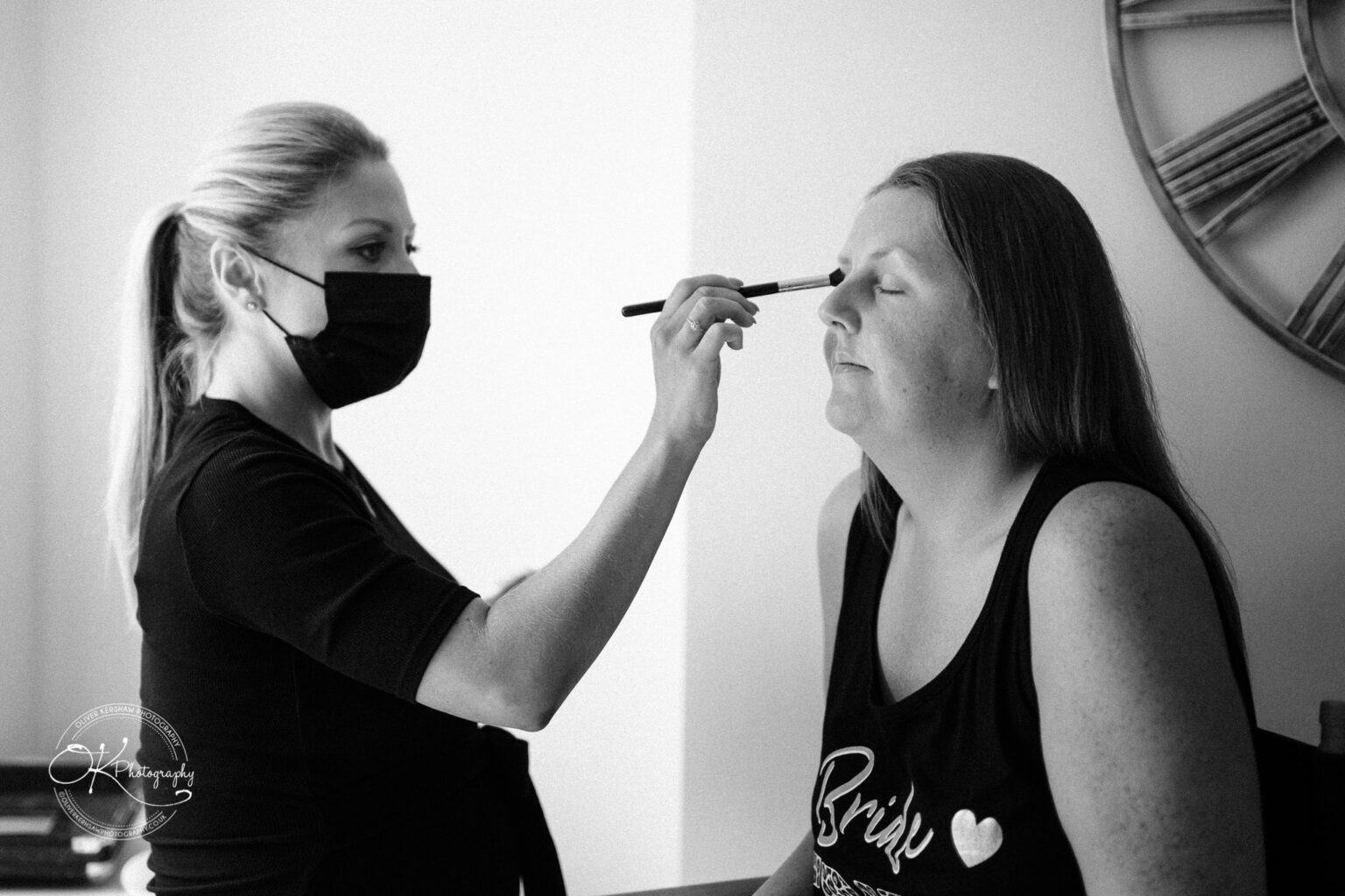 A makeup artist, wearing a face mask, applies makeup to a seated woman with her eyes closed.