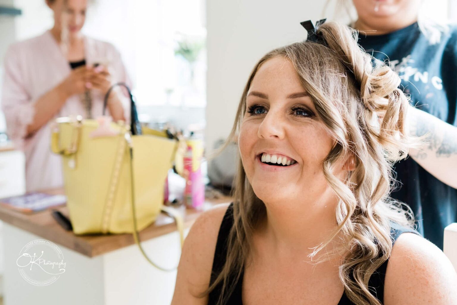 A woman with curled hair is smiling while another person styles her hair. In the background, another woman is using a mobile phone next to a yellow handbag on a counter.
