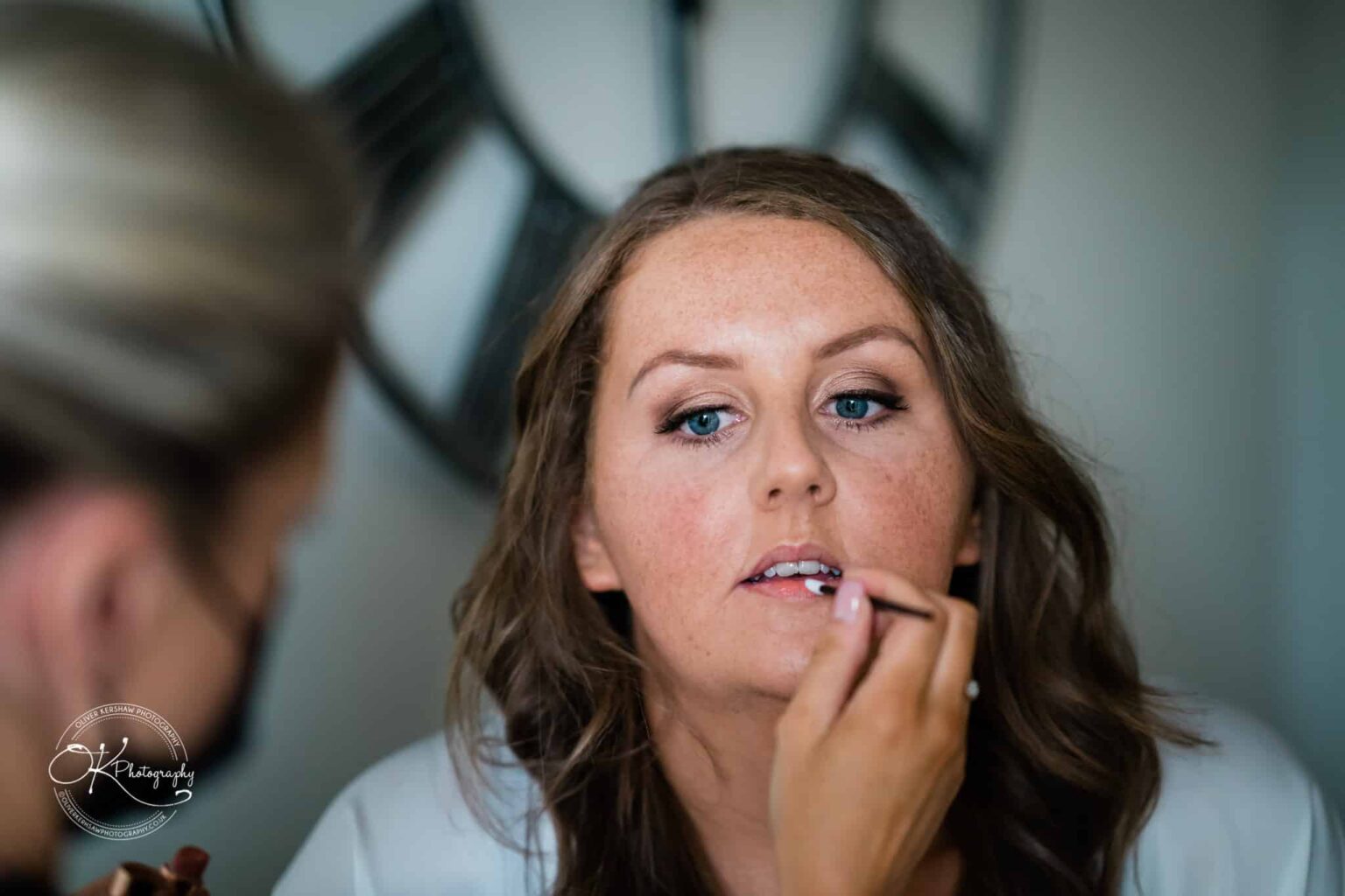 Close-up of a woman having her makeup applied by another person, focusing on her lips, with a blurred clock in the background.