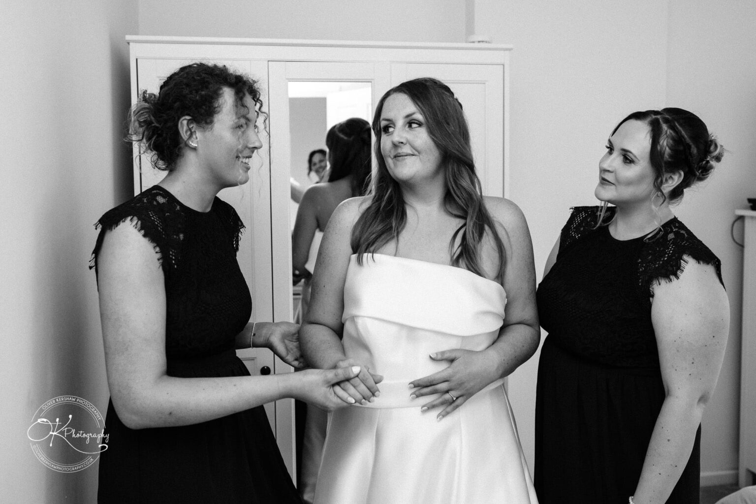 Three women in conversation, with one in a white bridal gown and the other two in black dresses, photographed in a room with a wardrobe in the background.