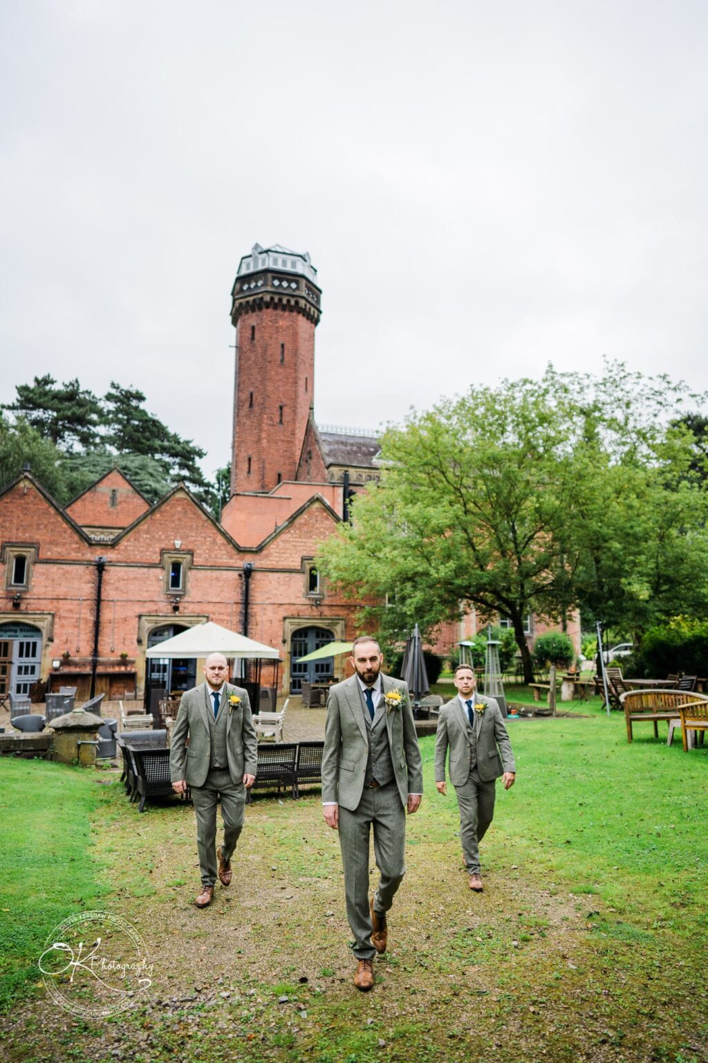 Three men in suits walking on a pathway with a historic brick building and tall tower in the background.