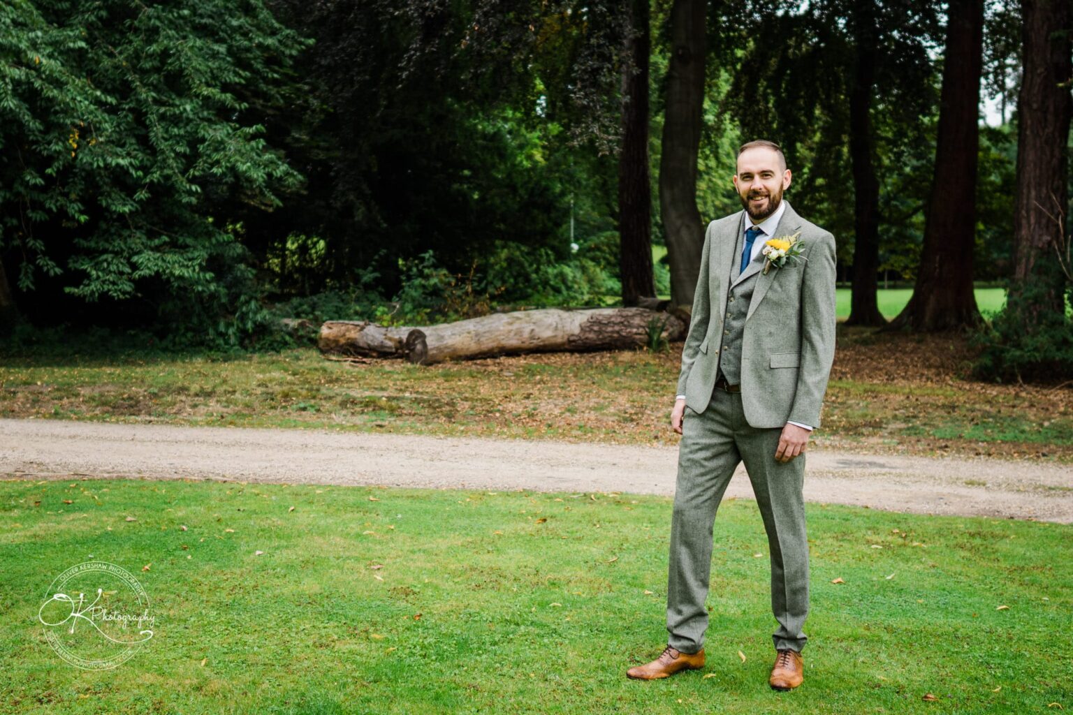 A man in a grey suit with a boutonniere, standing on grass with trees and a fallen log in the background.