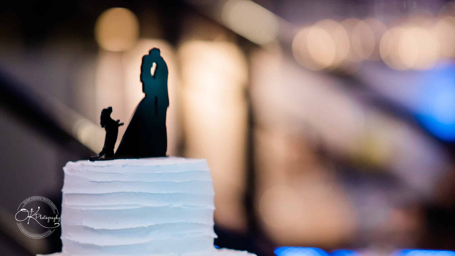 Close-up of a wedding cake topper depicting a bride and groom in a romantic pose, positioned on a white frosted cake with a bokeh background.