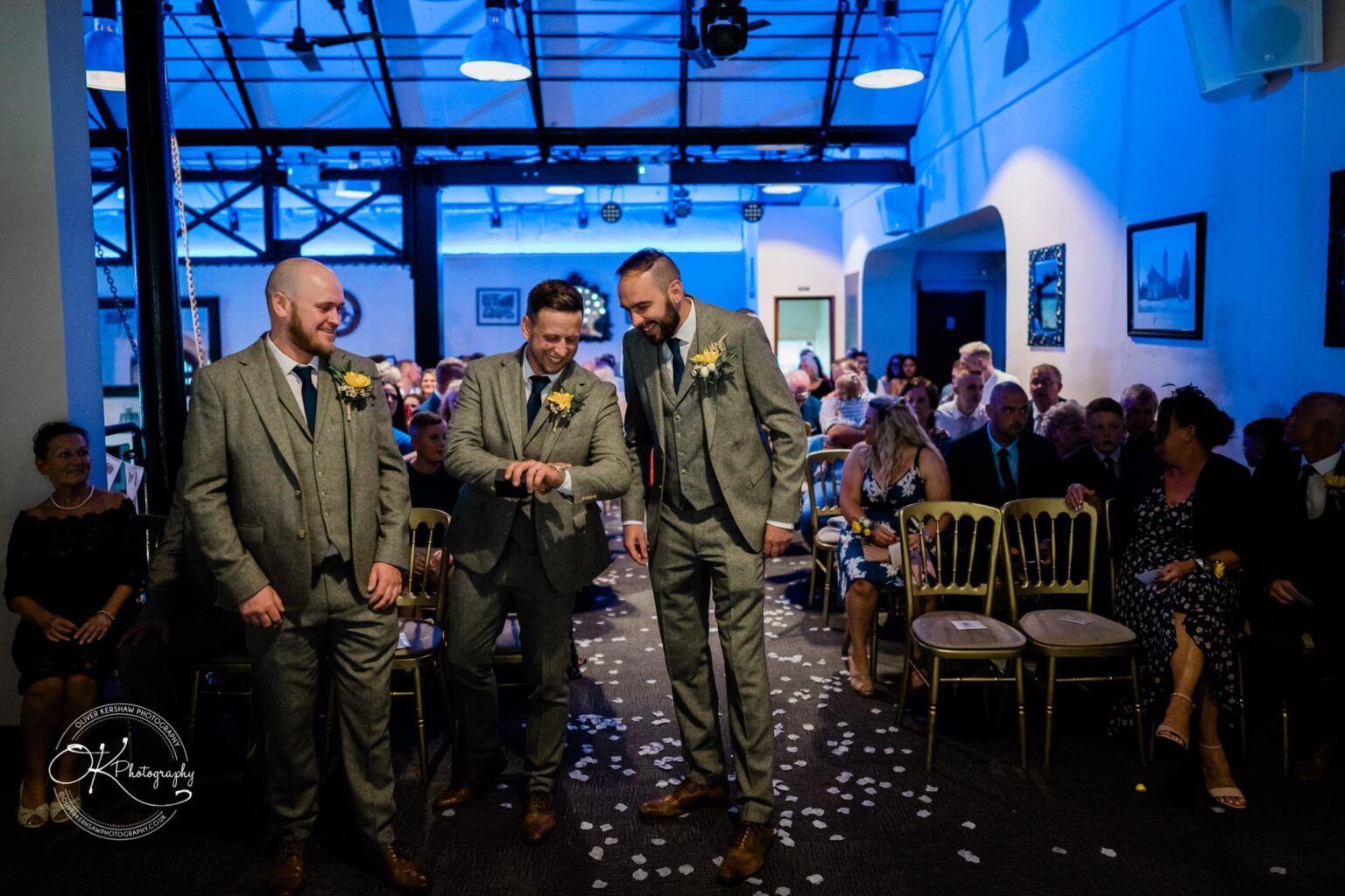 Three men in grey suits and yellow boutonnieres standing at the front of an indoor wedding venue, with guests seated behind them in rows.