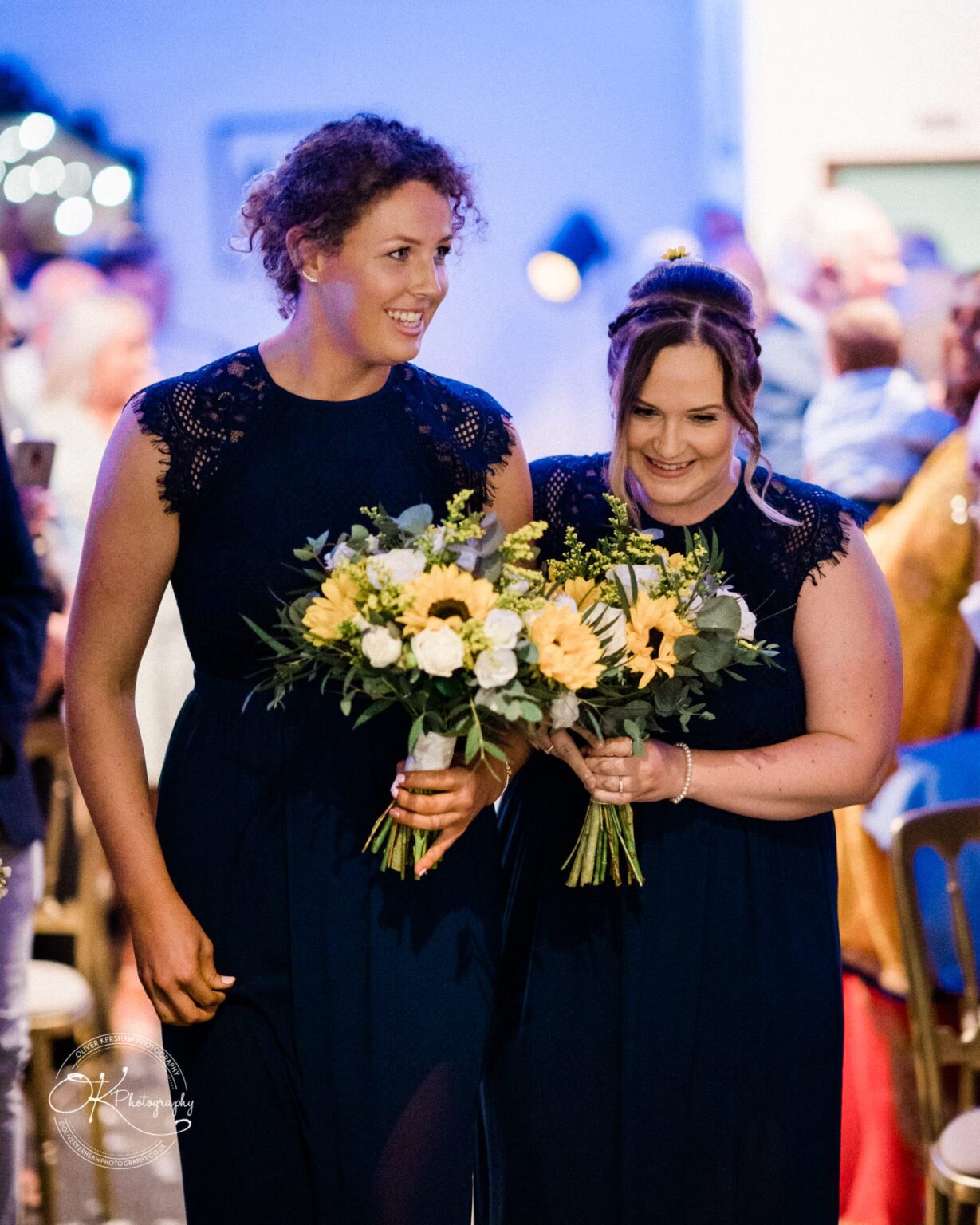 Two women in navy blue dresses holding bouquets of yellow and white flowers, smiling and standing close together at a formal event.