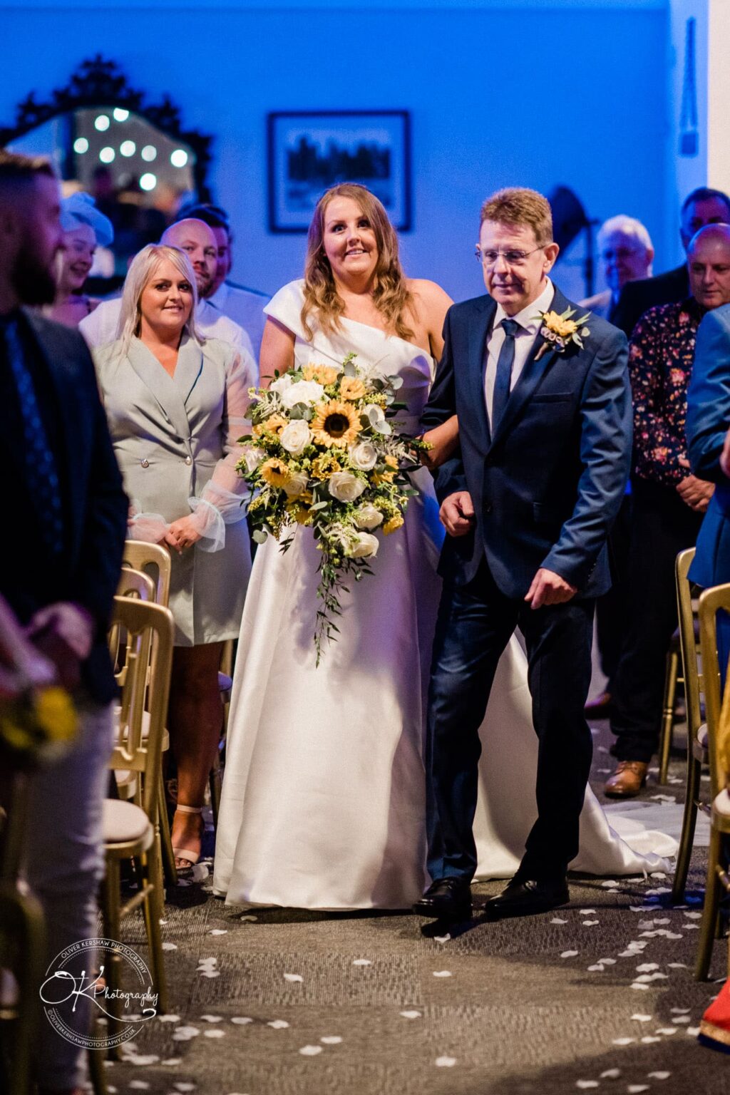 Bride walking down the aisle with her father at a wedding ceremony, holding a bouquet of sunflowers and white roses, surrounded by guests in a dimly lit venue.