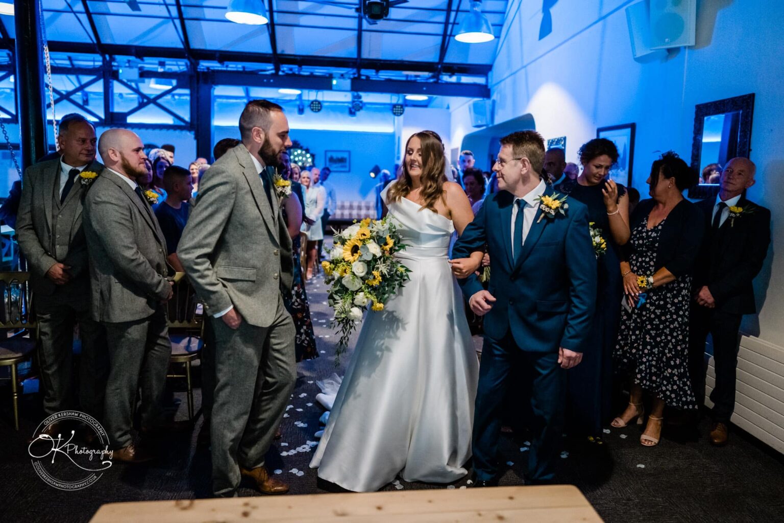 A bride in a white dress walks down the aisle with a man in a blue suit, surrounded by the wedding party and guests in a well-lit indoor venue.