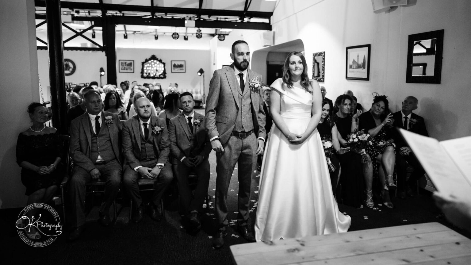 A bride and groom stand at the front of a room during a wedding ceremony, with guests seated behind them in rows. The black and white photo captures the moment seriously and intimately.