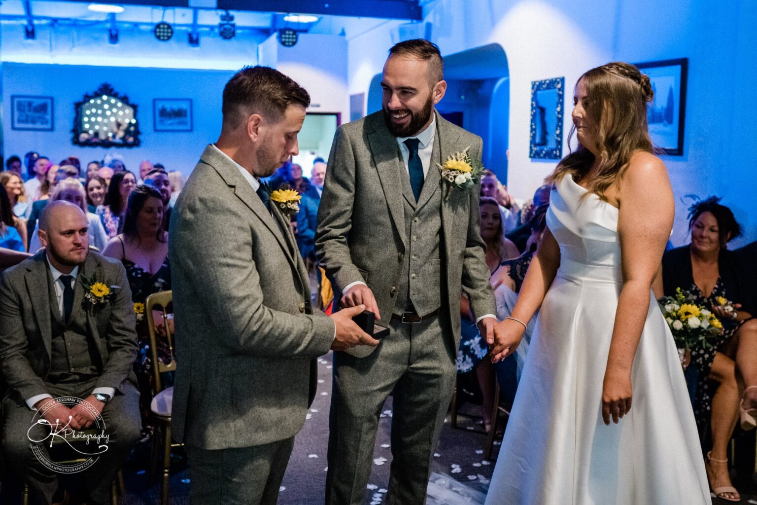 A wedding ceremony with a bride and groom holding hands, the groom smiling, and a groomsman presenting the rings. The audience is seated in the background.