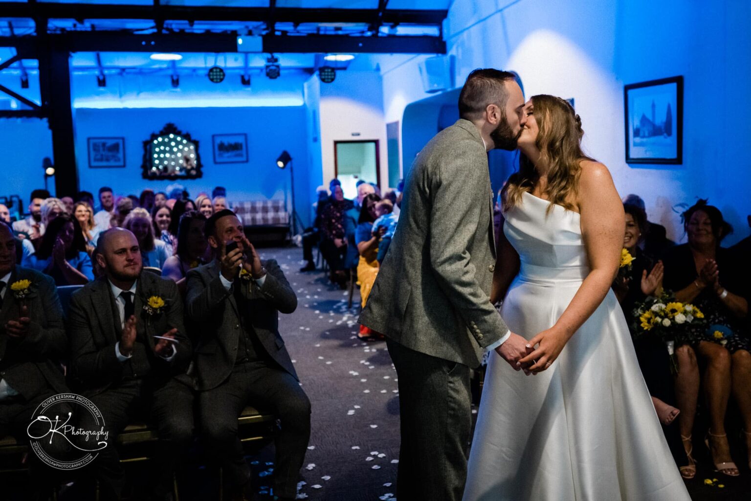 A bride and groom kiss in front of an applauding audience at a wedding ceremony in a dimly lit room.