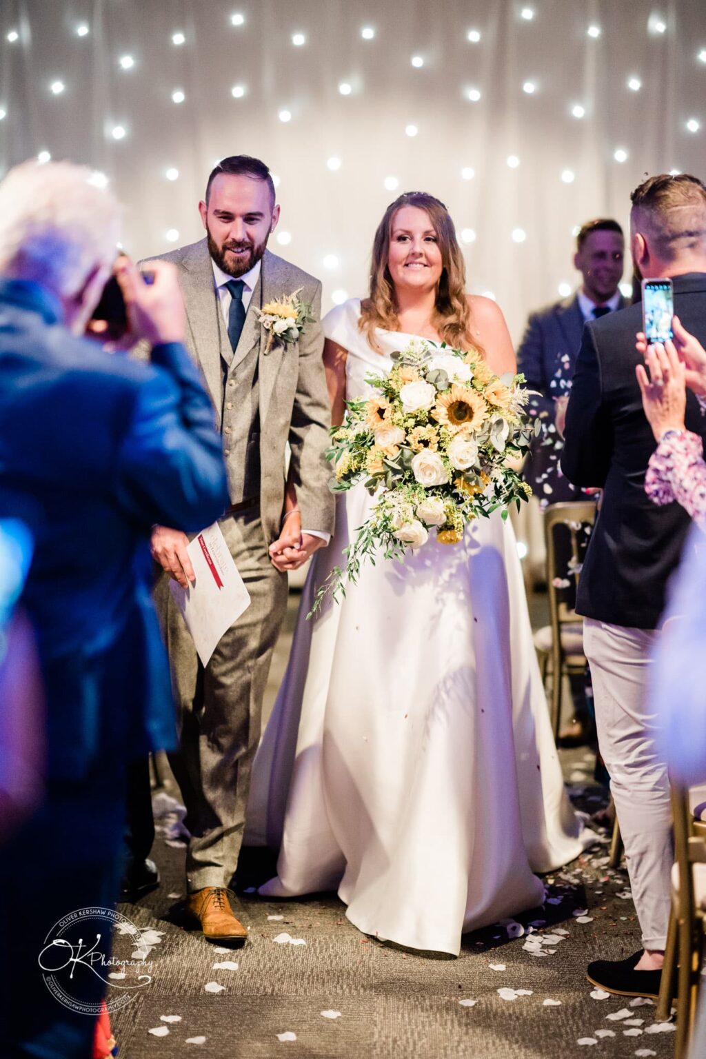 Newlywed couple holding hands and smiling while walking down the aisle, surrounded by guests taking photos and recording the moment, with a backdrop of glowing fairy lights.