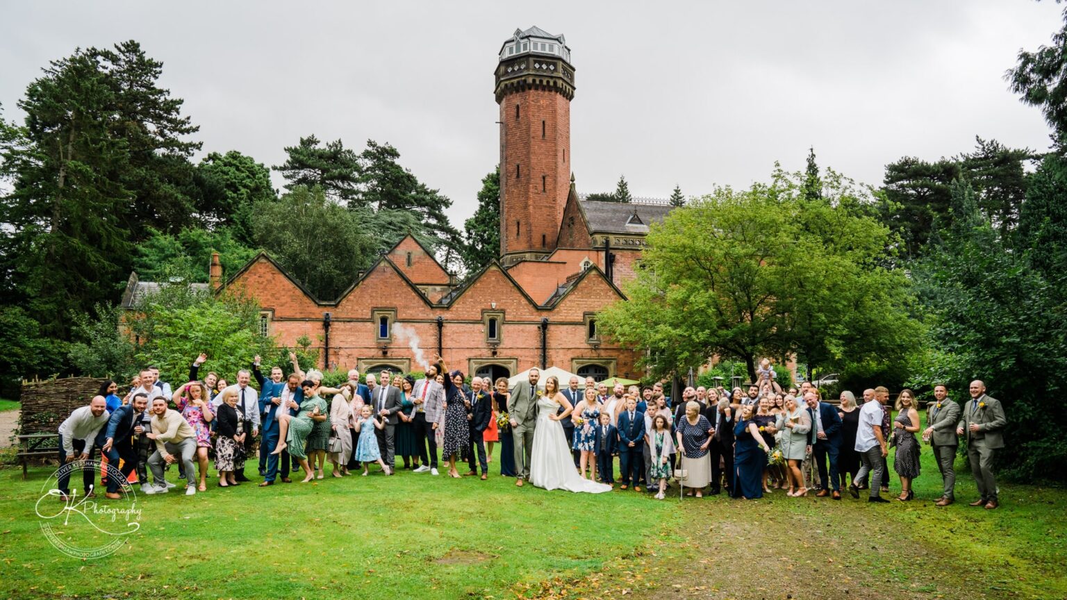 A large group of wedding guests standing and celebrating in front of a historic red brick building with a tall tower, surrounded by greenery.
