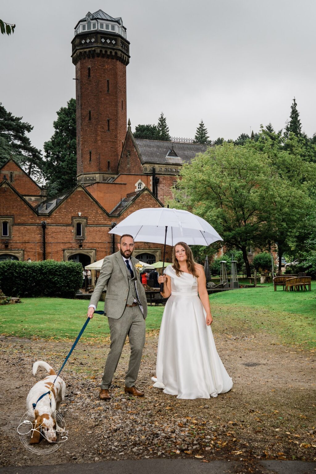 A bride and groom standing outside a historic brick building with a tower, holding an umbrella and a dog on a leash.