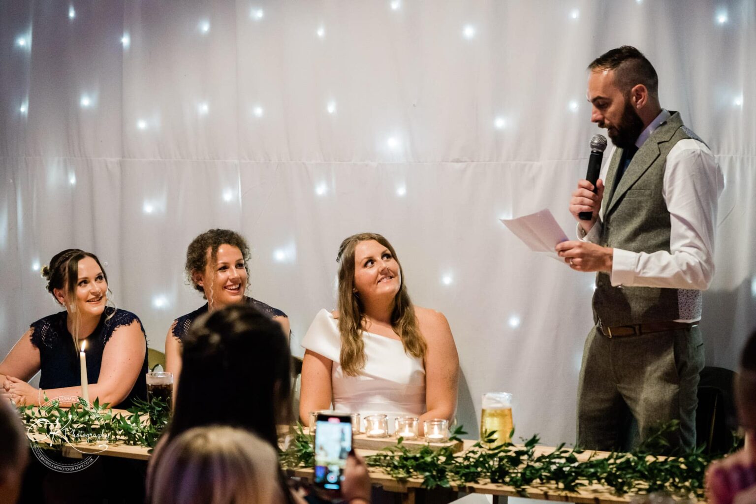 A man in a suit delivers a speech with a microphone while three women, including a bride in a white dress, sit at a decorated table and listen, smiling.