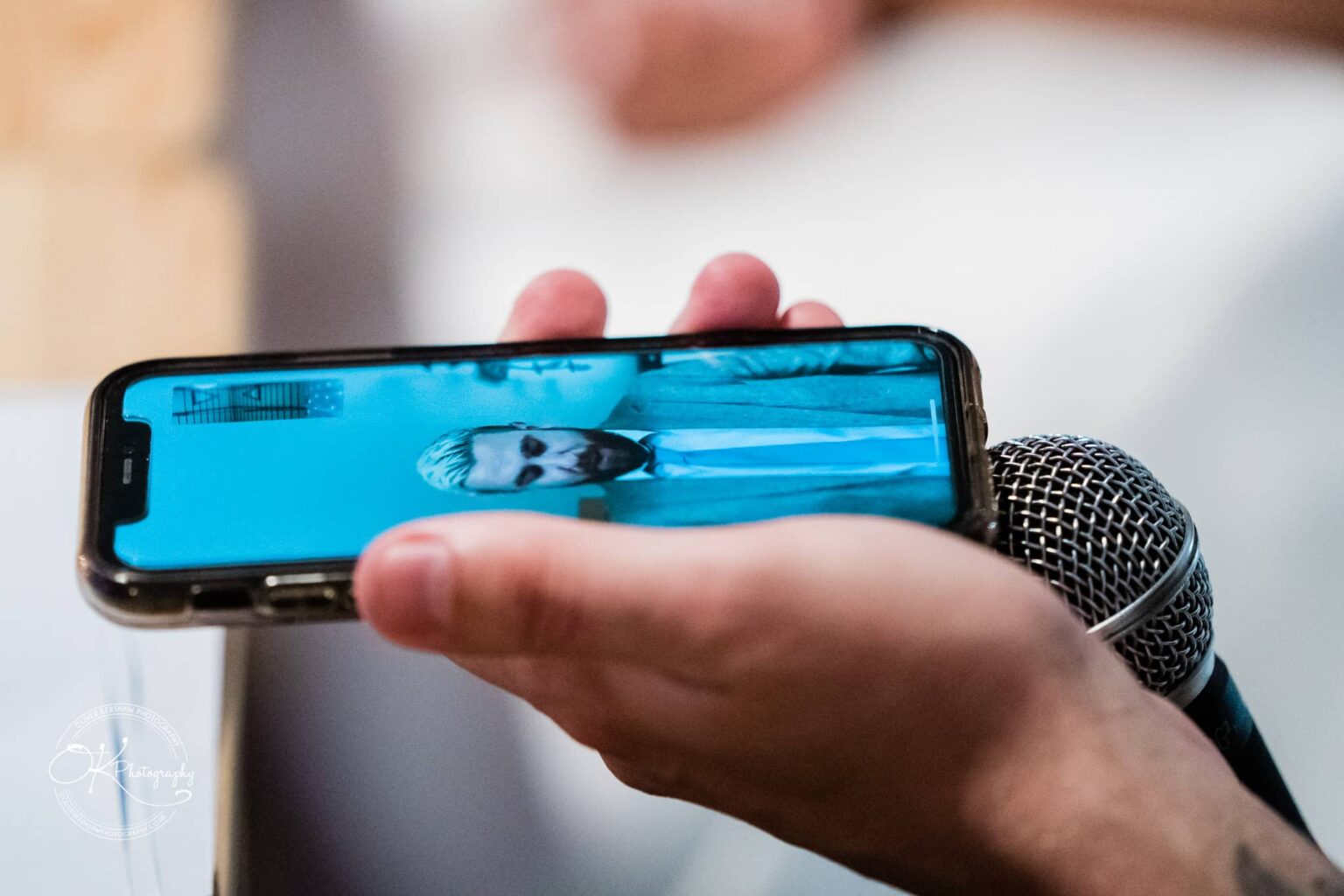 A close-up of a hand holding a smartphone displaying an image of a person in a suit, with a microphone held near the phone.