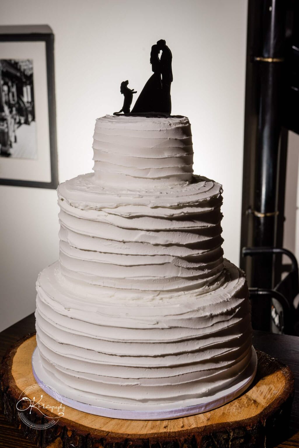 Three-tiered white wedding cake with rustic textured icing, topped with a silhouette topper of a couple and a dog, placed on a wooden log cake stand.