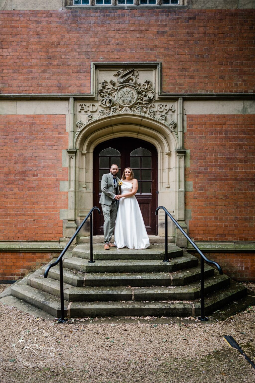 A newlywed couple stands on steps in front of an arched door of a historic brick building with ornate stonework and the inscription "AD 1894" above.