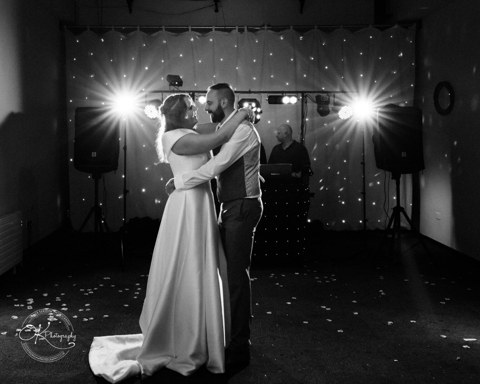 A couple dancing at their wedding with a DJ in the background, illuminated by bright lights.