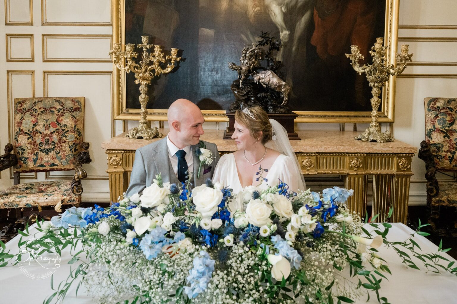 A bride and groom sitting at a table adorned with blue and white flowers, in an ornate room with antique decor at Warwick Castle.