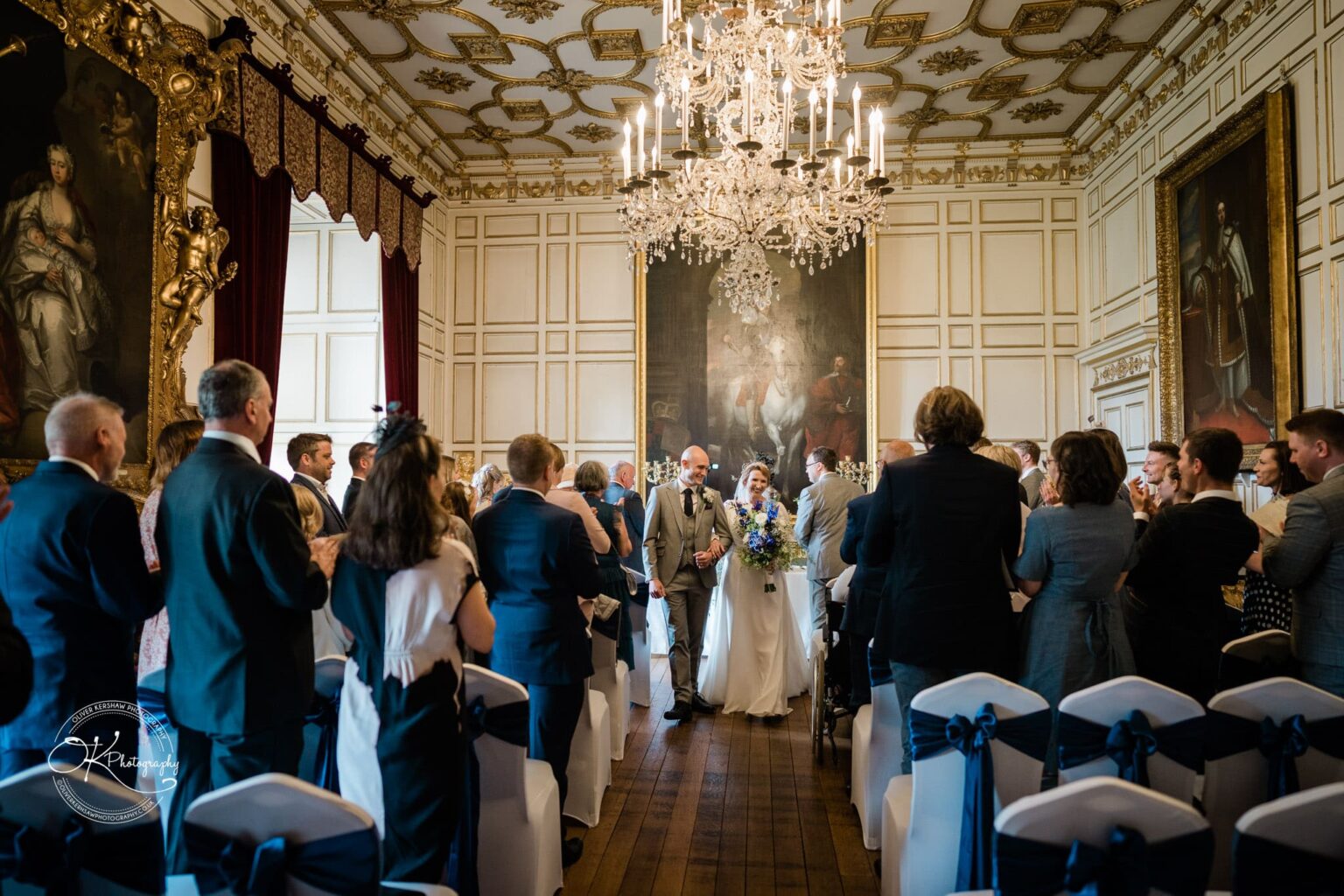 A wedding ceremony in a grand hall with chandeliers, ornate ceiling details, and large paintings on the walls. Guests are seated and standing, applauding as the newlywed couple walks down the aisle.