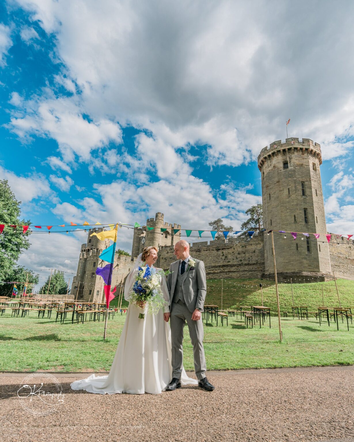A bride and groom stand together in the foreground with Warwick Castle in the background, decorated with colourful bunting on a sunny day.